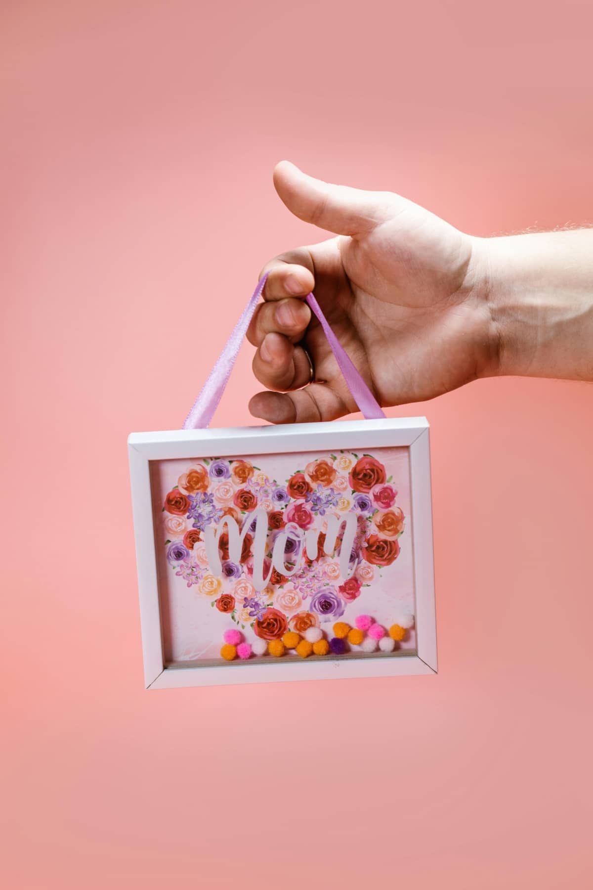 Close-up of hand holding a floral 'Mom' shadow box against a pink background, perfect gift for Mother's Day.